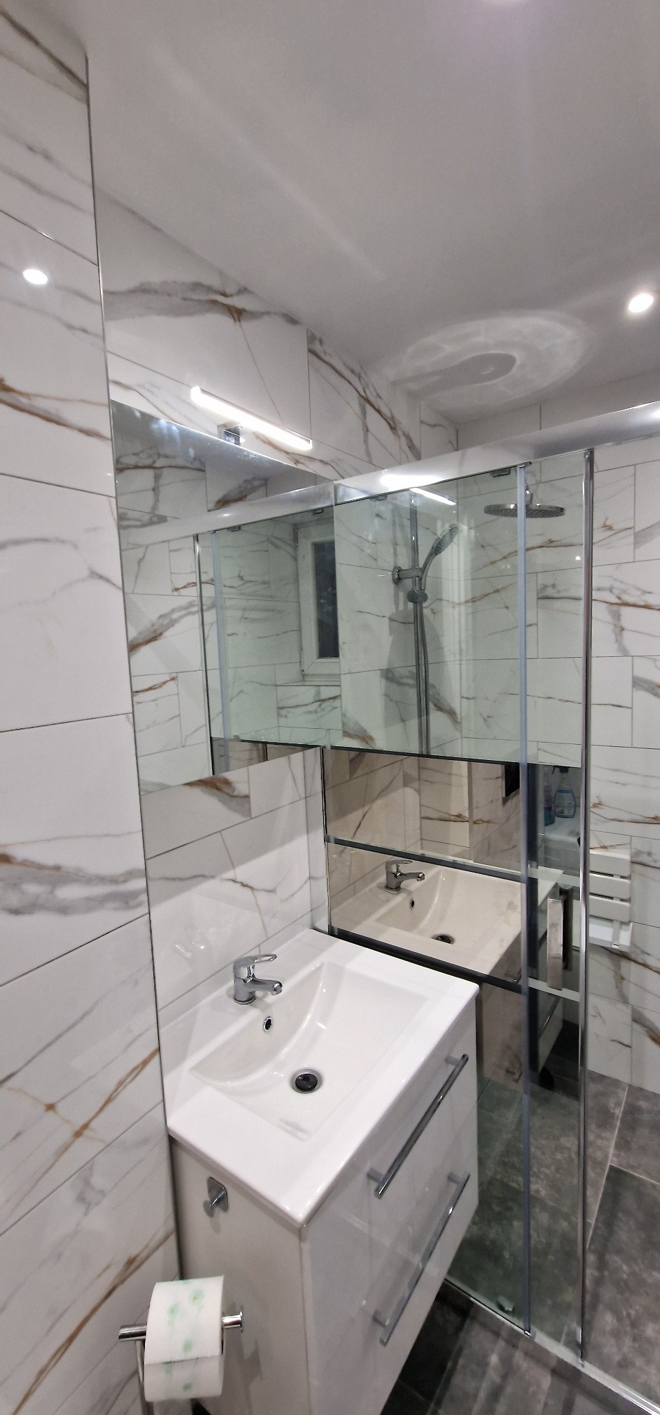 Nowoczesna łazienka z białymi płytkami imitującymi marmur, kabina prysznicowa ze szklanymi drzwiami i biała umywalka z szafką, widoczne odbicie w lustrze