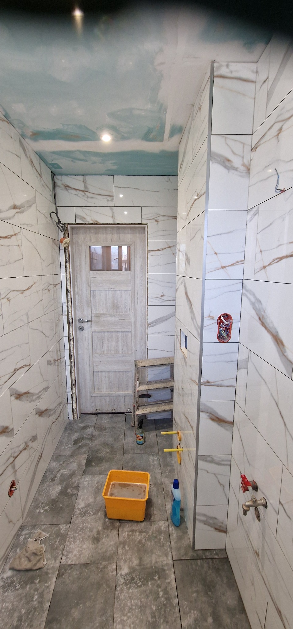 Wnętrze łazienki w trakcie wykańczania: białe płytki imitujące marmur na ścianach, szare płytki podłogowe, sufit z efektem nieba, widoczne instalacje hydrauliczne i elektryczne, drabina, wiadro...