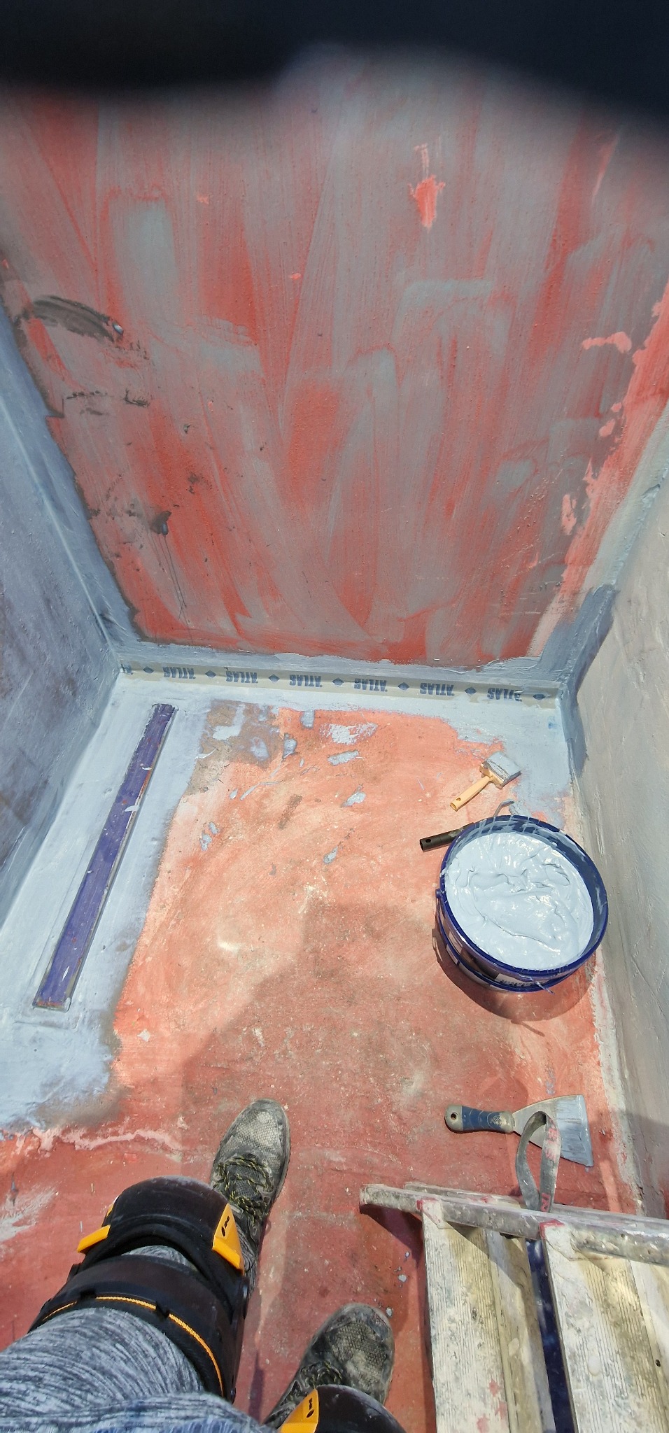 Przygotowanie podłoża w kabinie prysznicowej: czerwona hydroizolacja z widocznymi pociągnięciami pędzla na ścianach i podłodze, taśma uszczelniająca ATLAS na łączeniach, wiadro z szarą masą...