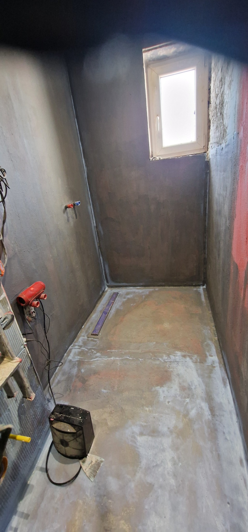 Wnętrze małej łazienki w trakcie przygotowań do remontu, ściany pokryte szarą warstwą hydroizolacji, widoczne przyłącza wodne, grzejnik elektryczny, drabina i łatka miernicza na podłodze.