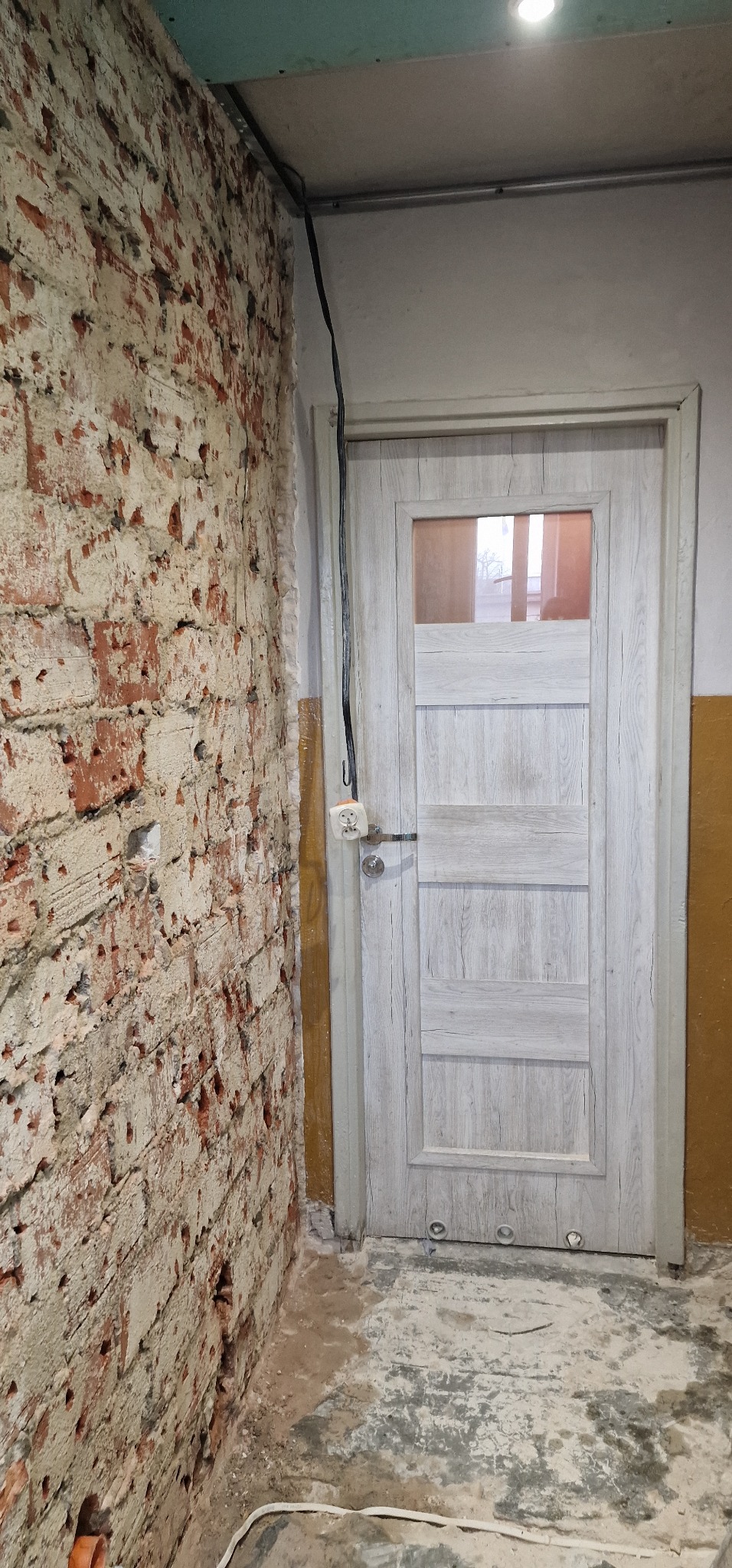 Nowe, jasne drzwi wewnętrzne z szybą, zamontowane w trakcie remontu w Głubczycach, widoczne na tle surowej, ceglanej ściany i odsłoniętej instalacji elektrycznej.