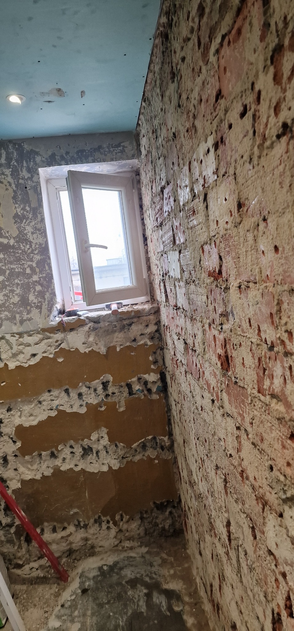 Fragment wnętrza w trakcie prac przygotowawczych: odsłonięta cegła na ścianie, resztki starego tynku, częściowo otwarte białe okno, surowy sufit z płyt gipsowych.