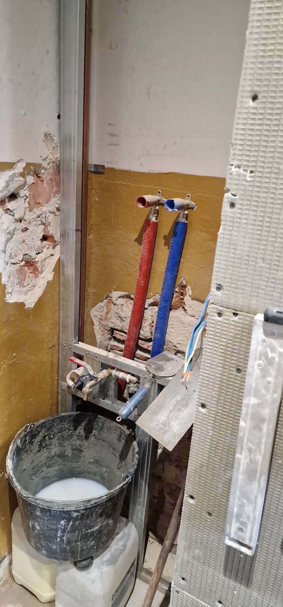 Wnętrze łazienki w trakcie remontu: widoczne rury ciepłej i zimnej wody, metalowy stelaż, wiadro z zaprawą, narzędzia murarskie i płyta kartonowo-gipsowa przygotowana do montażu.