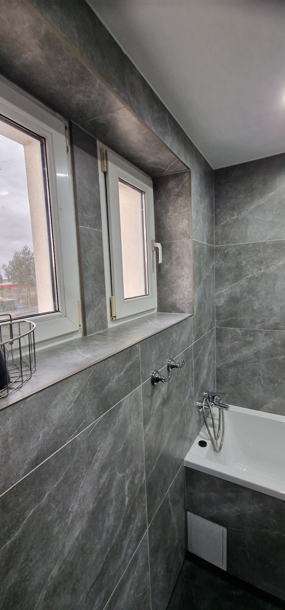 Wykończona łazienka z szarymi płytkami imitującymi marmur na ścianach, biała wanna z chromowaną baterią, dwa okna z białymi ramami i metalowy koszyk na blacie pod oknami.