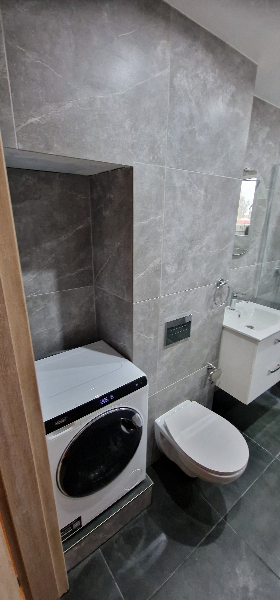 Nowoczesna łazienka z szarymi płytkami imitującymi marmur, białą pralką Haier umieszczoną we wnęce, wiszącą toaletą i umywalką z szafką.