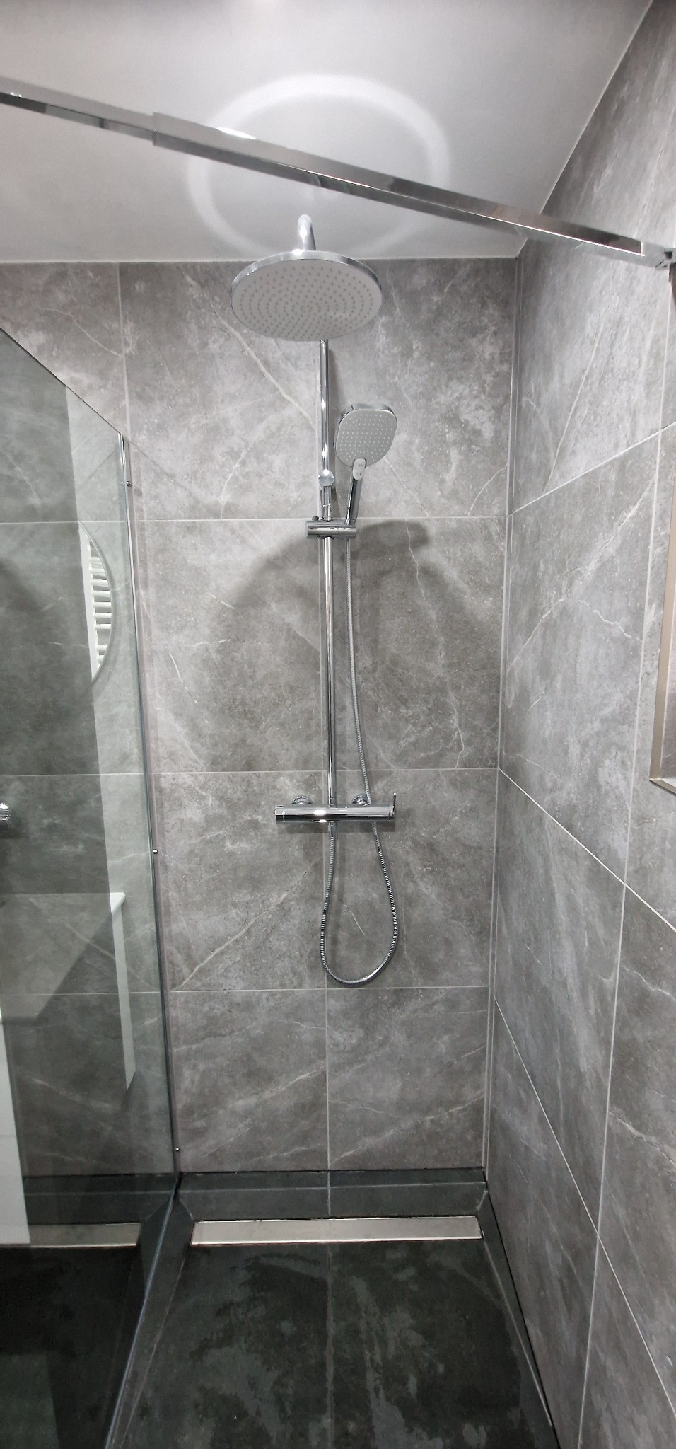 Nowoczesna kabina prysznicowa z deszczownicą i szarymi płytkami imitującymi marmur, czarny brodzik z odpływem liniowym, szklana ściana kabiny