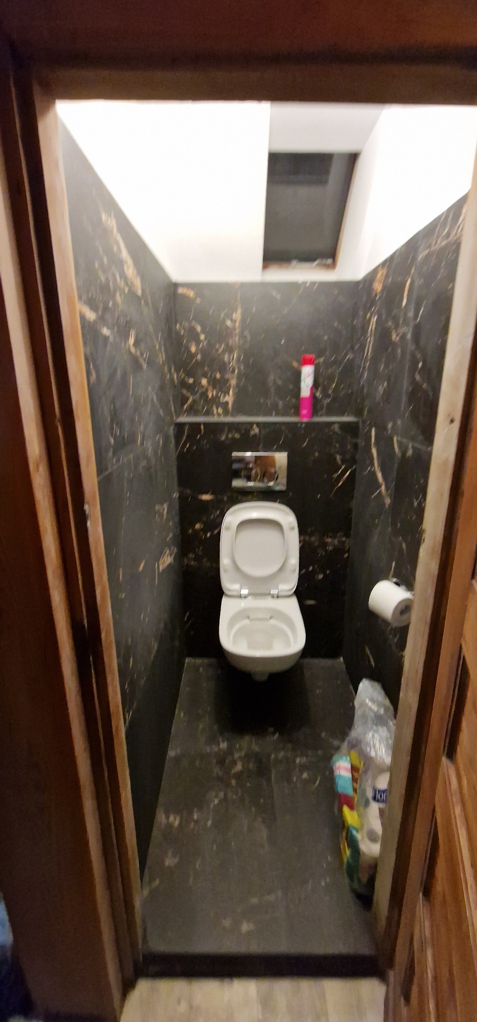Wnętrze małej łazienki wykończonej czarnym marmurem z białymi żyłami, widoczna toaleta z otwartą klapą, rolka papieru toaletowego i zapas papieru w folii, oświetlenie sufitowe w górnej części kadru.