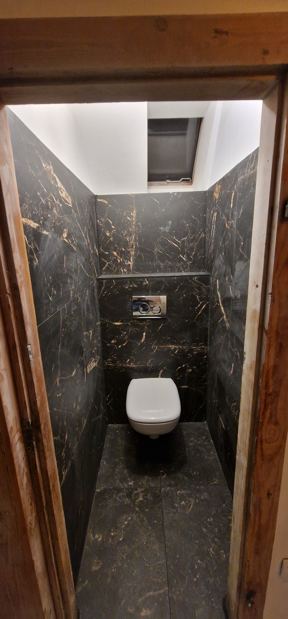 Mała toaleta wykończona czarnymi płytkami imitującymi marmur ze złotymi żyłami, widok zza drewnianej ościeżnicy, na suficie okno dachowe.