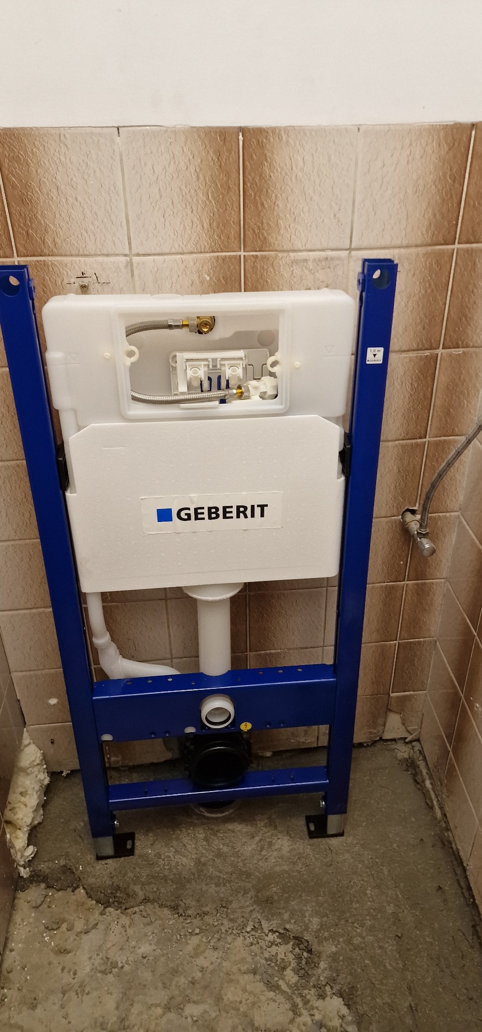 Stelaż podtynkowy Geberit Duofix zamontowany w trakcie prac wykończeniowych w łazience, widoczne przyłącza i odpływ, tło stanowią stare, brązowe płytki.