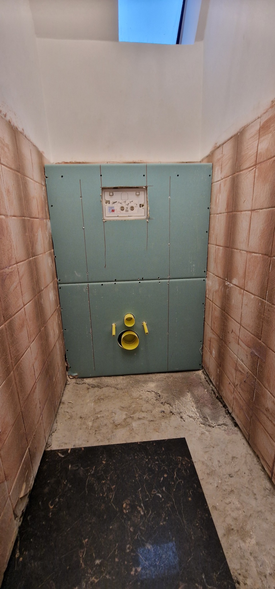 Przygotowanie do montażu podtynkowego stelaża WC na płycie kartonowo-gipsowej w łazience z brązową glazurą, widoczne żółte rury kanalizacyjne i fragment czarnej podłogi.