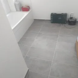 Świeżo wyremontowana łazienka z szarymi płytkami podłogowymi, białą wanną i skrzynką narzędziową w rogu. Widoczny fragment ściany z fakturą.