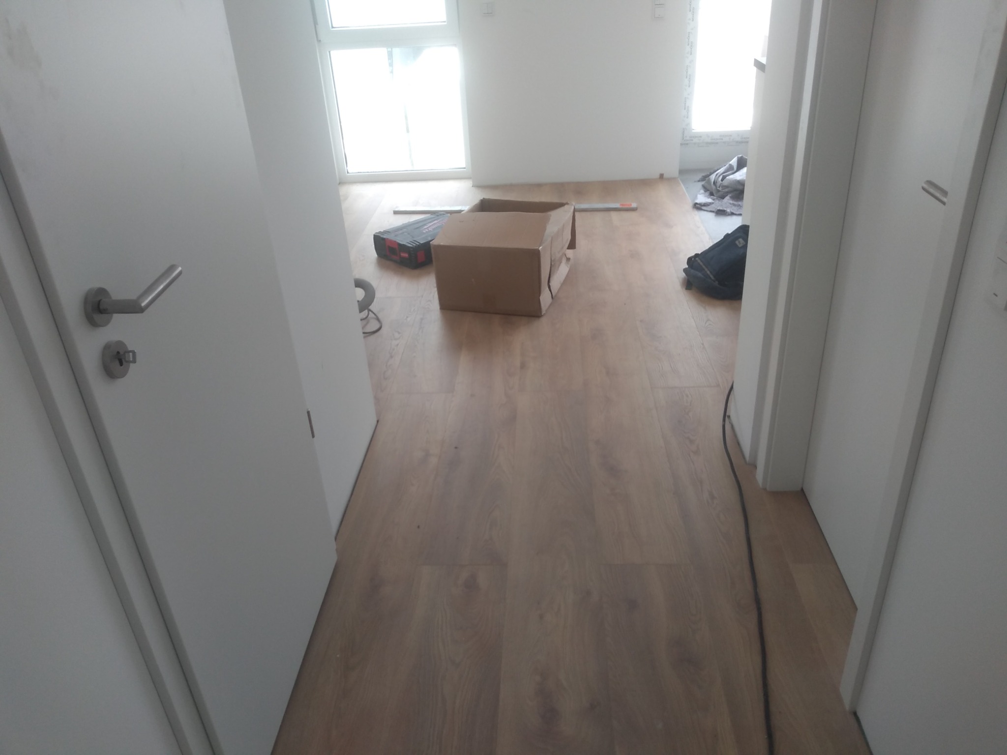 Wnętrze pokoju w trakcie remontu: panele podłogowe, białe ściany, narzędzia i karton na podłodze, widok z korytarza z otwartymi drzwiami.