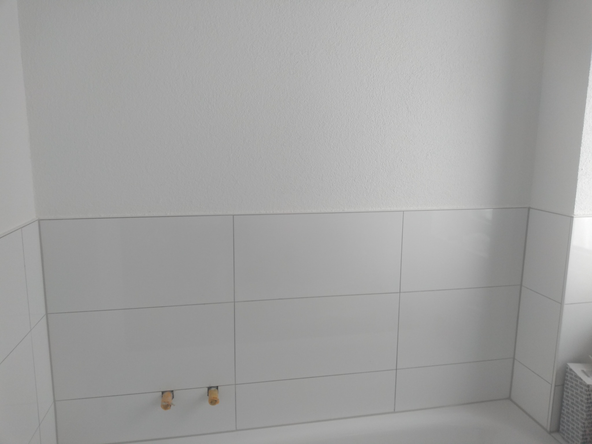 Świeżo ułożone, białe, prostokątne płytki ścienne w łazience, z widocznymi wystającymi rurami instalacji wodnej.