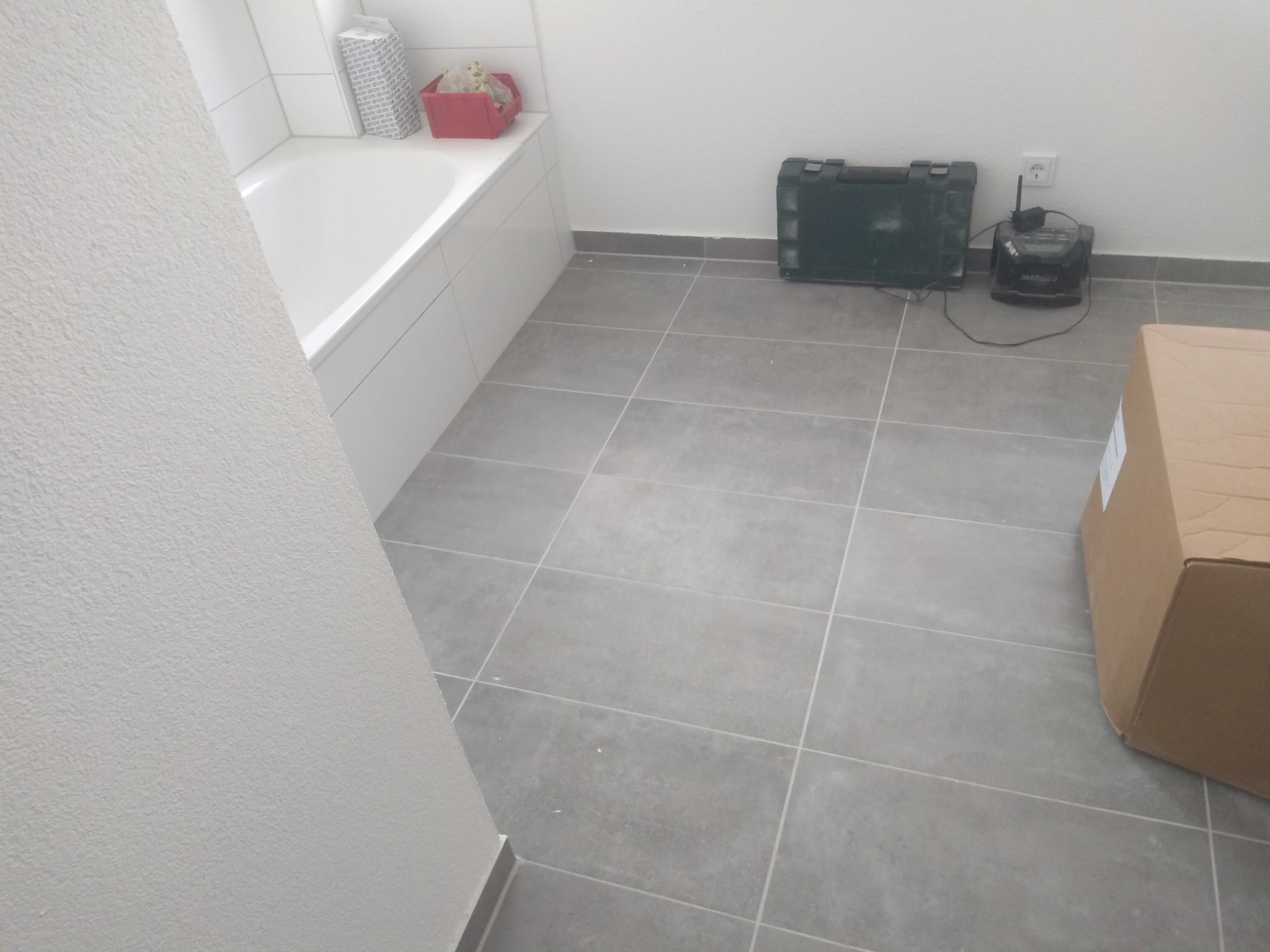 Świeżo wyremontowana łazienka z szarymi płytkami podłogowymi, białą wanną i skrzynką narzędziową w rogu. Widoczny fragment ściany z fakturą.