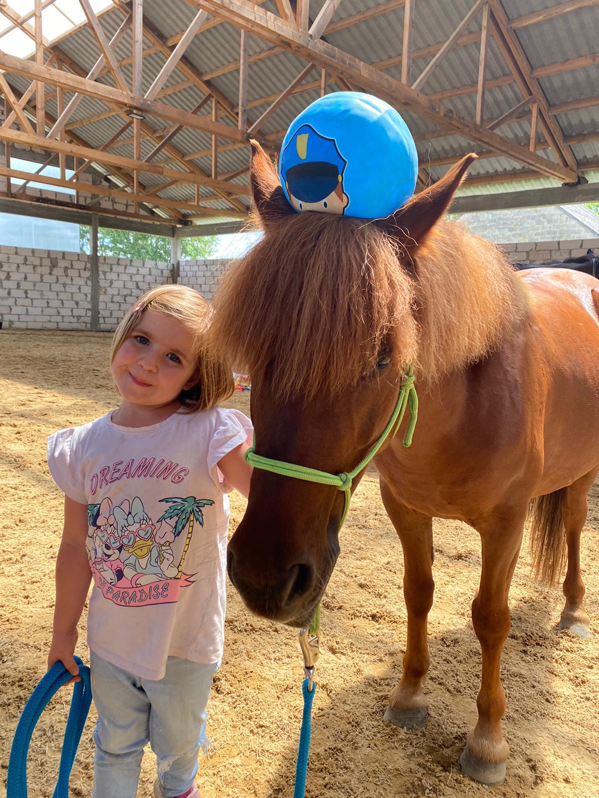 Dziewczynka z blond włosami w koszulce z motywem Disneya, stojąca obok brązowego konia z pluszową kulą policjanta na głowie, w jasnym, zadaszonym maneżu.