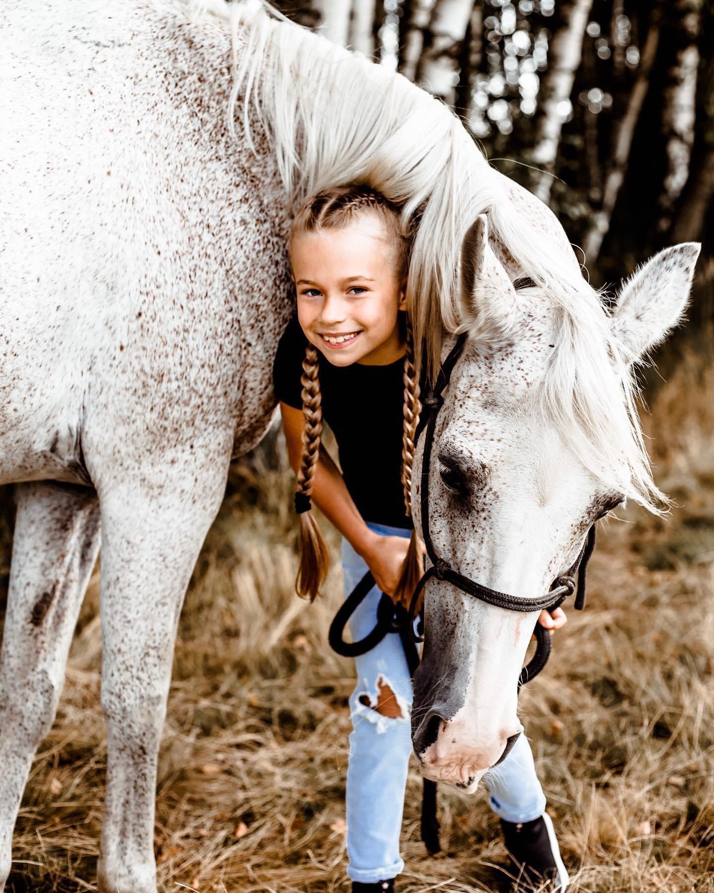 Uśmiechnięta dziewczynka z blond warkoczami przytula się do siwego konia w brązowe plamki, trzymając go za uzdę na tle suchych traw i drzew.