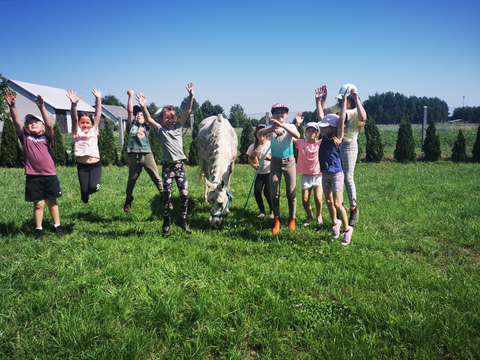 Grupa dzieci skacze z radości na zielonej łące obok białego konia, część z nich ma pomalowane twarze, w tle widoczny budynek i drzewa.