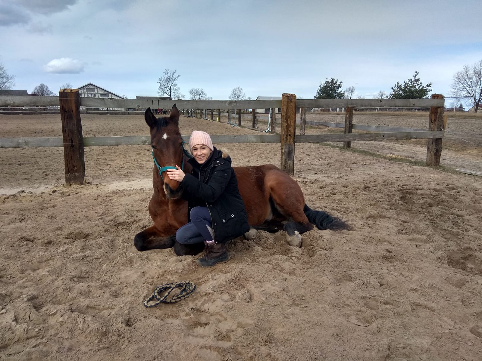 Kobieta w różowej czapce kuca obok leżącego brązowego konia z białą gwiazdą na czole, na piaszczystym padoku z drewnianym ogrodzeniem w tle.