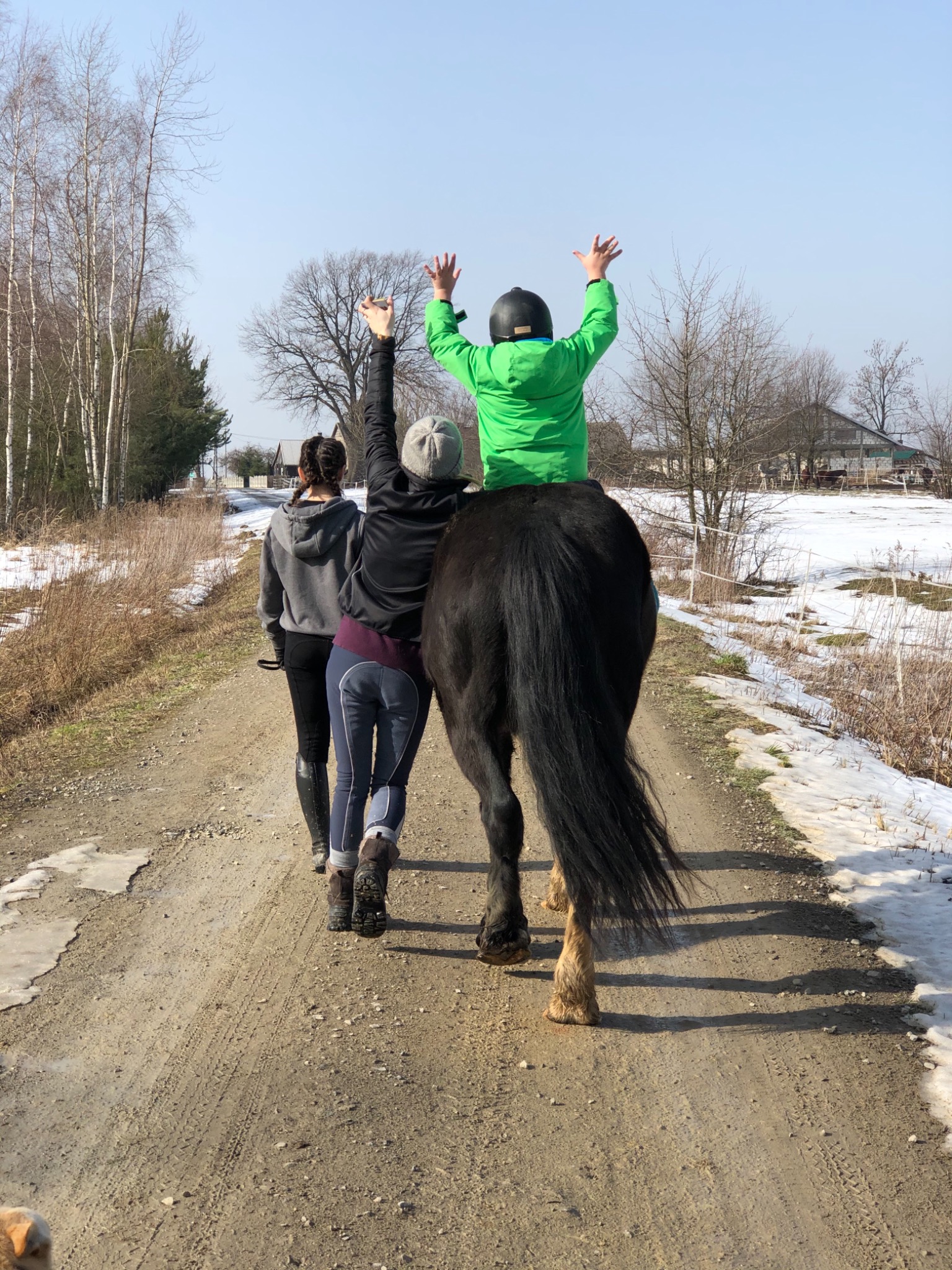 Trzy osoby i koń na polnej drodze; dziecko w zielonej kurtce siedzi na koniu z podniesionymi rękami, dwie kobiety idą obok, w tle zabudowania i resztki śniegu.