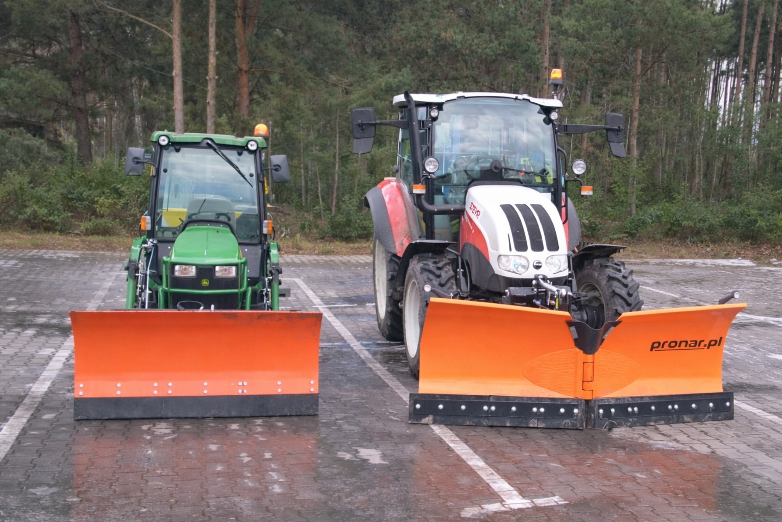 Dwa traktory z pomarańczowymi pługami do odśnieżania, stojące na parkingu z widocznym lasem w tle. Jeden traktor jest zielony, drugi biało-czerwony.