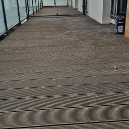 Sokólscy - Nowoczesny balkon z drewnianą podłogą kompozytową i szklanymi balustradami, widoczny głośnik Makita, pochmurne niebo w tle.