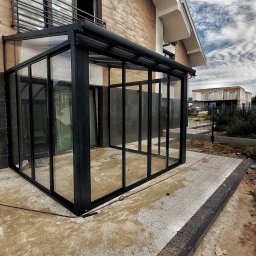 Sokólscy - Zadaszona, przeszklona konstrukcja balkonu z czarnymi profilami, widoczna z zewnątrz na tle budynku z cegły i nieba z chmurami.