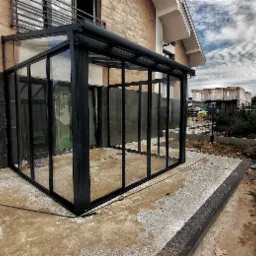 Zadaszona, przeszklona konstrukcja balkonu z czarnymi profilami, widoczna z zewnątrz na tle budynku z cegły i nieba z chmurami.