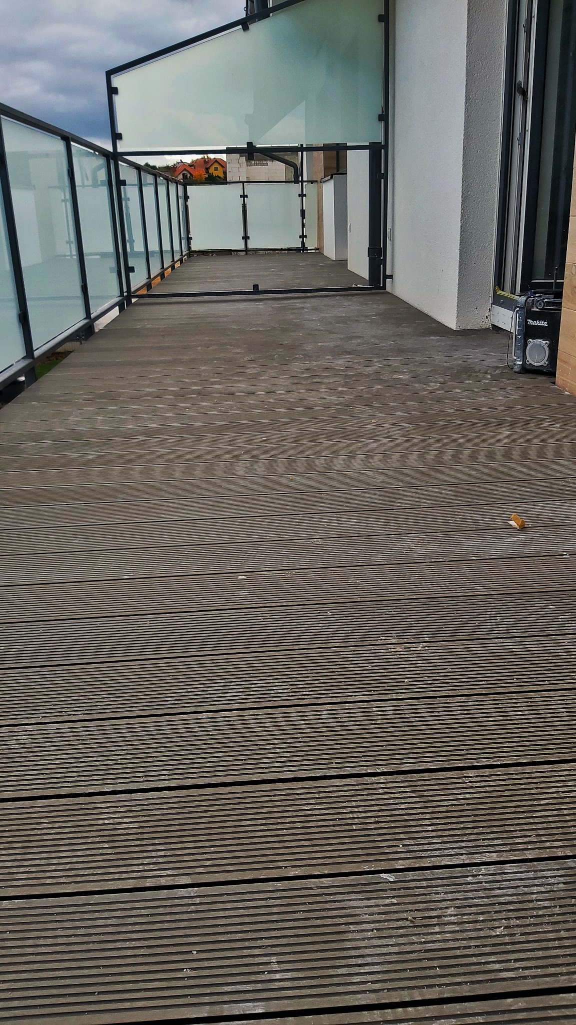 Nowoczesny balkon z drewnianą podłogą kompozytową i szklanymi balustradami, widoczny głośnik Makita, pochmurne niebo w tle.