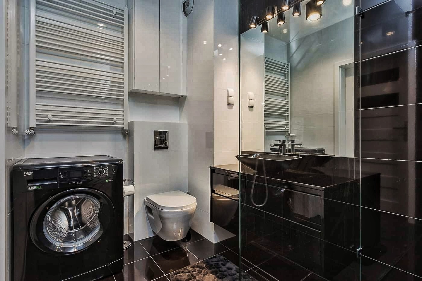 Nowoczesna łazienka z czarną pralką Beko, podwieszaną toaletą, grzejnikiem drabinkowym oraz czarną umywalką na szafce, odbijającą się w dużym lustrze z oświetleniem punktowym.