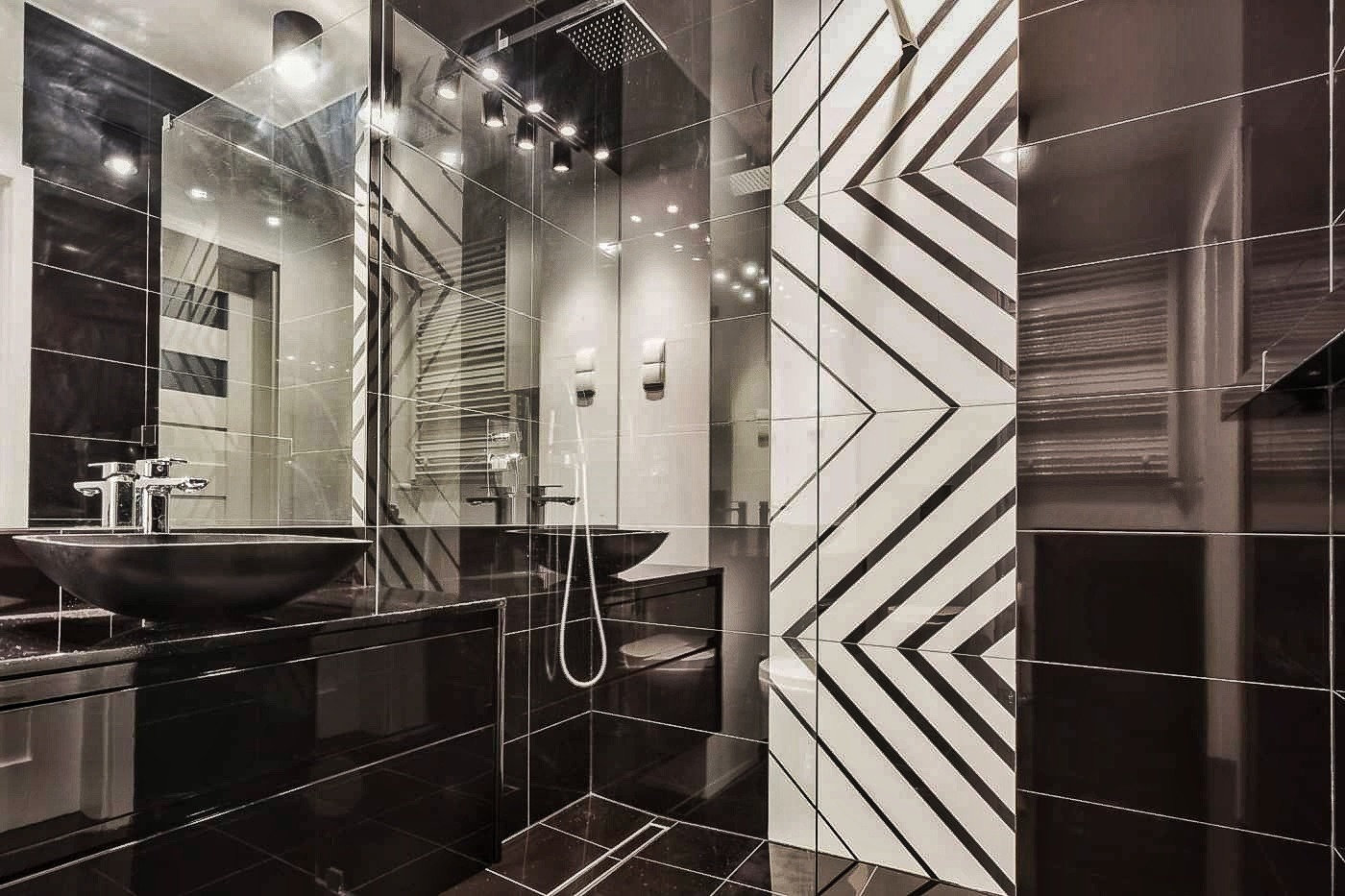 Nowoczesna łazienka z czarnymi błyszczącymi płytkami, umywalką w kształcie misy, szklanym panelem prysznicowym i ścianą z wzorem w zygzaki.