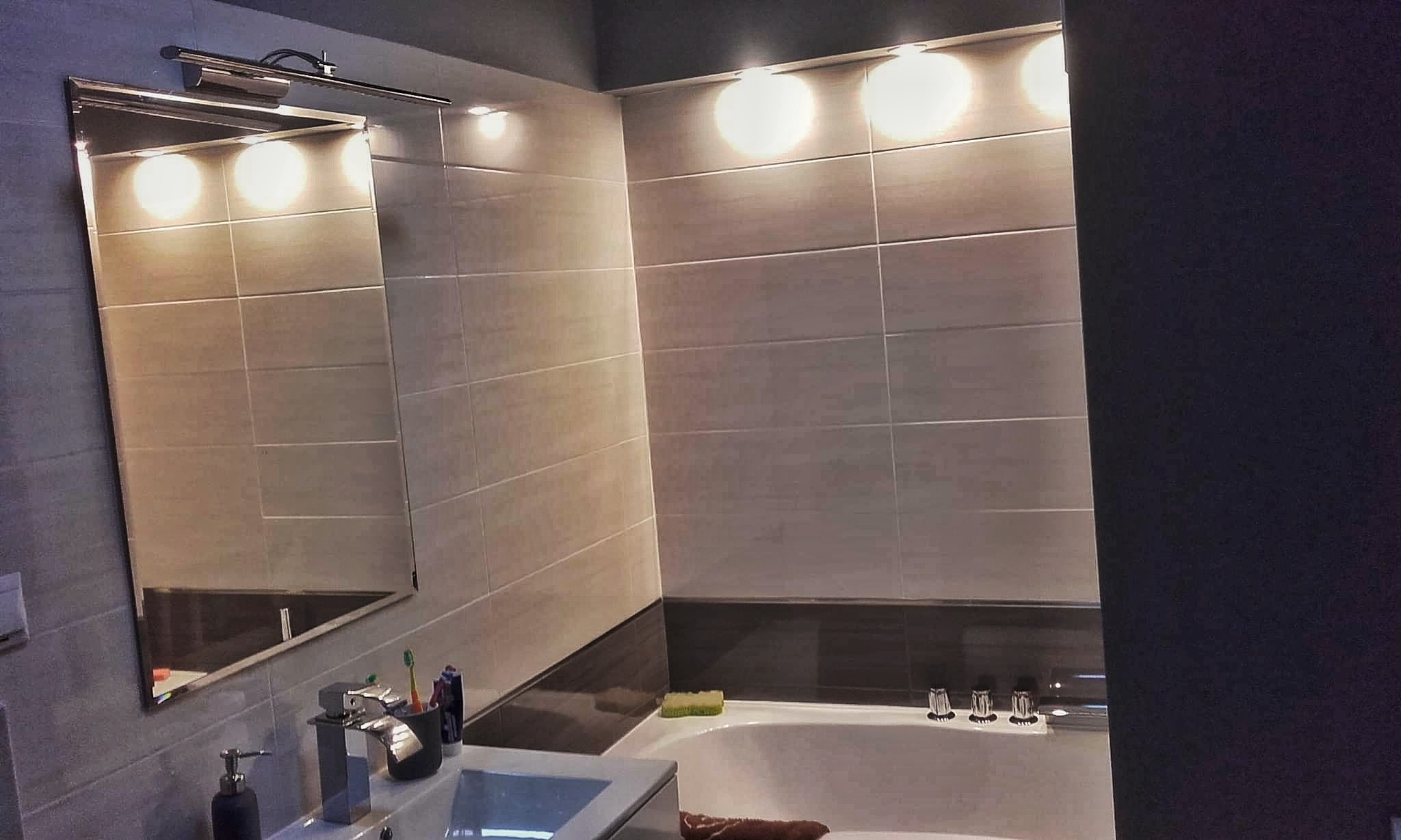 Wykończona łazienka w szarych płytkach z podświetlanym lustrem, widoczna umywalka z baterią, wanna i akcesoria łazienkowe.