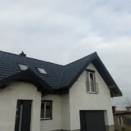 Nowoczesny dom jednorodzinny z ciemną dachówką ceramiczną, białymi ścianami i czarnymi detalami, widok z dołu na tle pochmurnego nieba.