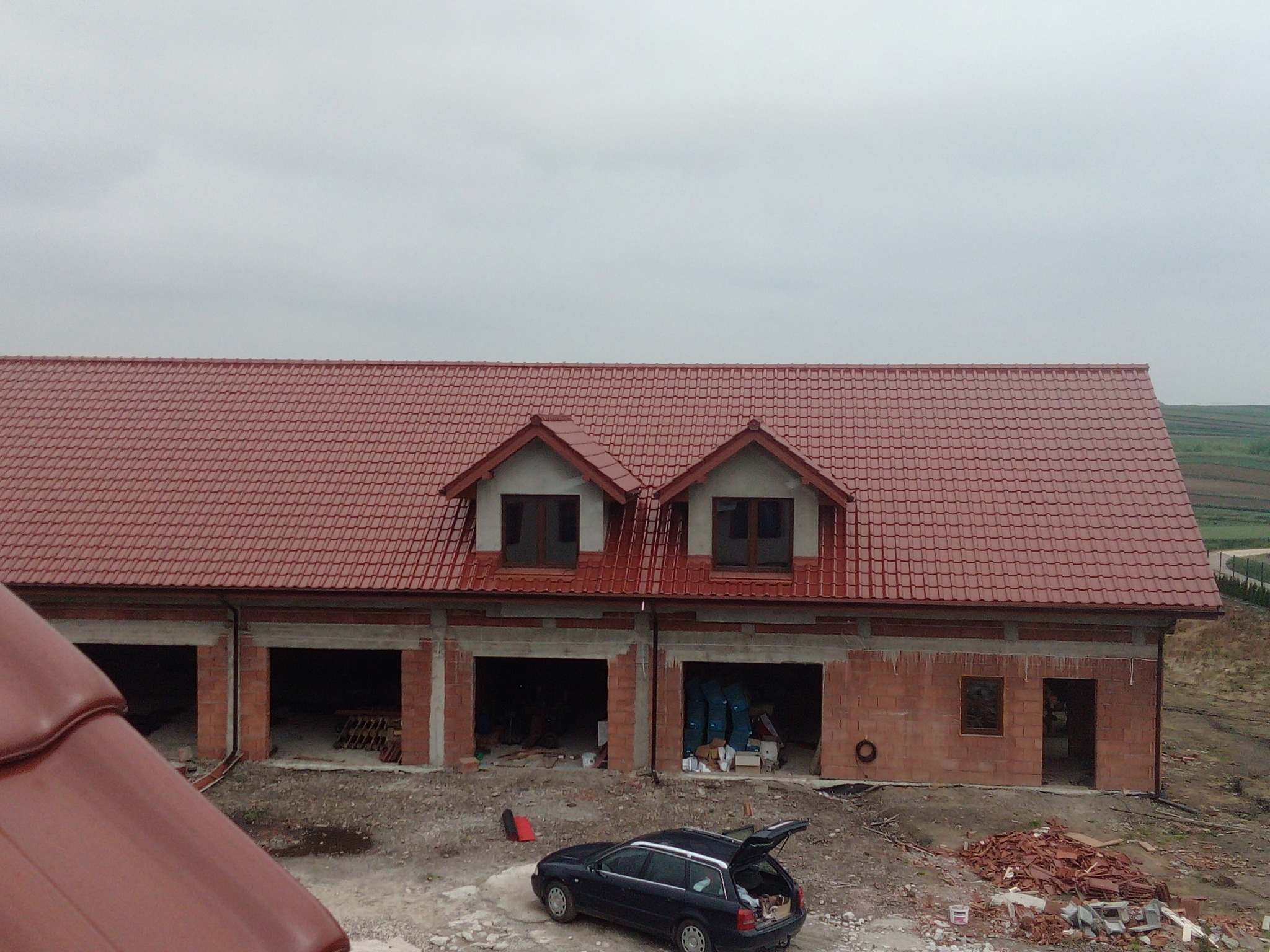 Nowy, czerwony dach z dachówki ceramicznej na budynku w trakcie budowy, widok z góry na budynek z otwartymi garażami, samochód kombi z otwartą klapą bagażnika na placu budowy.