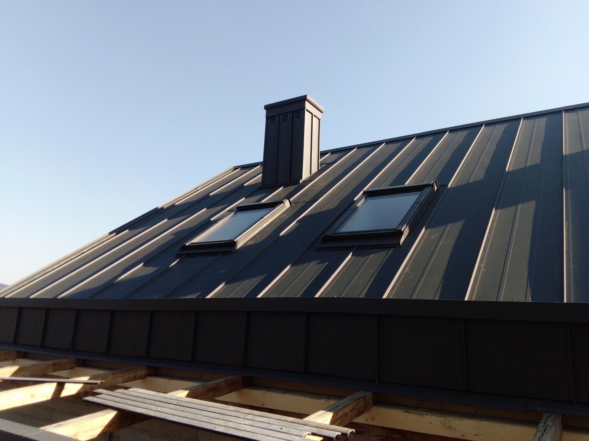 Nowoczesny, grafitowy dach z blachy trapezowej z dwoma oknami dachowymi i kominem, widoczny w trakcie montażu, z elementami drewnianej konstrukcji w dolnej części kadru.