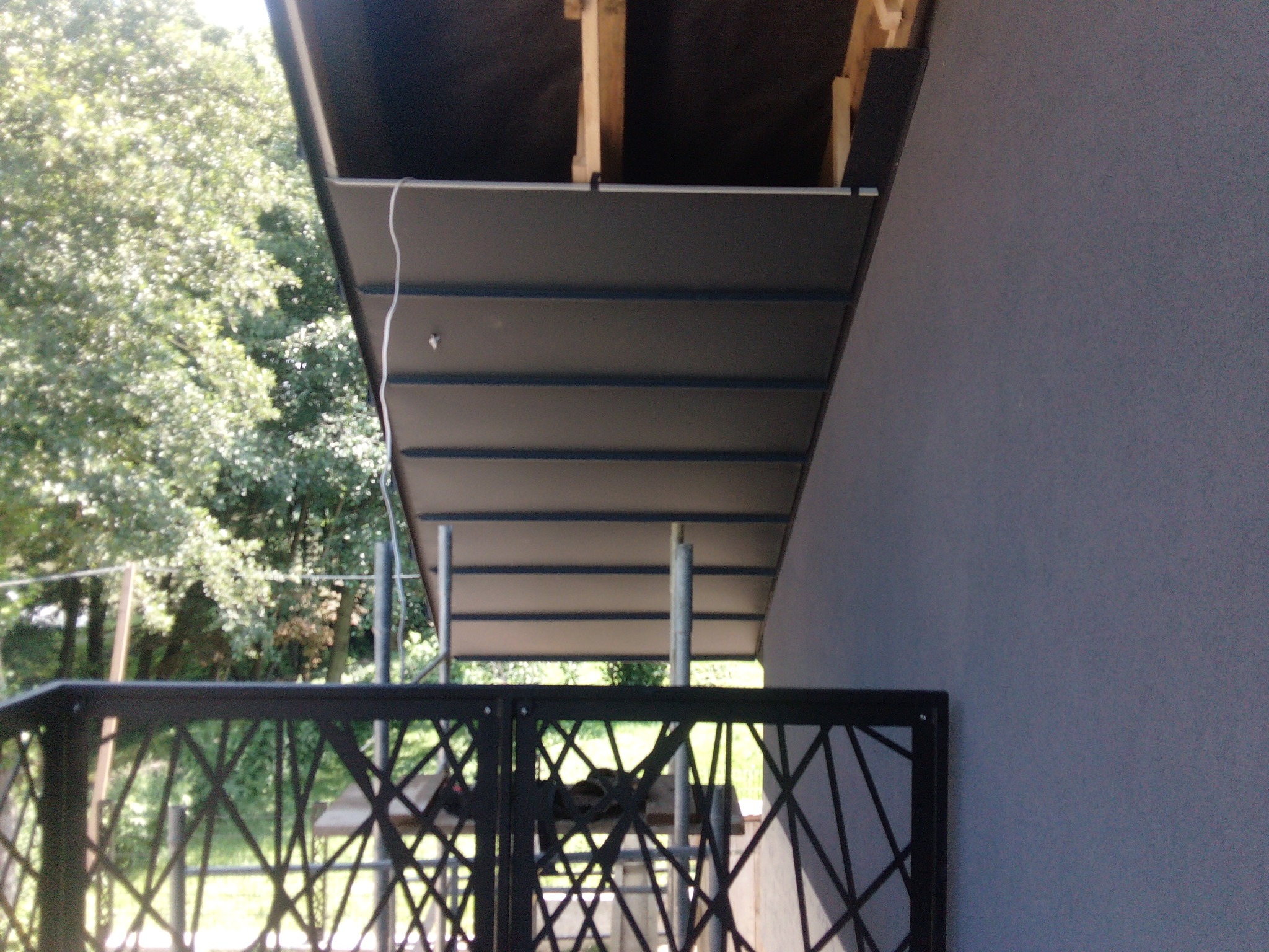 Montaż podbitki dachowej z widoczną konstrukcją drewnianą, metalowymi rusztowaniami i dekoracyjnym ogrodzeniem balkonowym w tle.