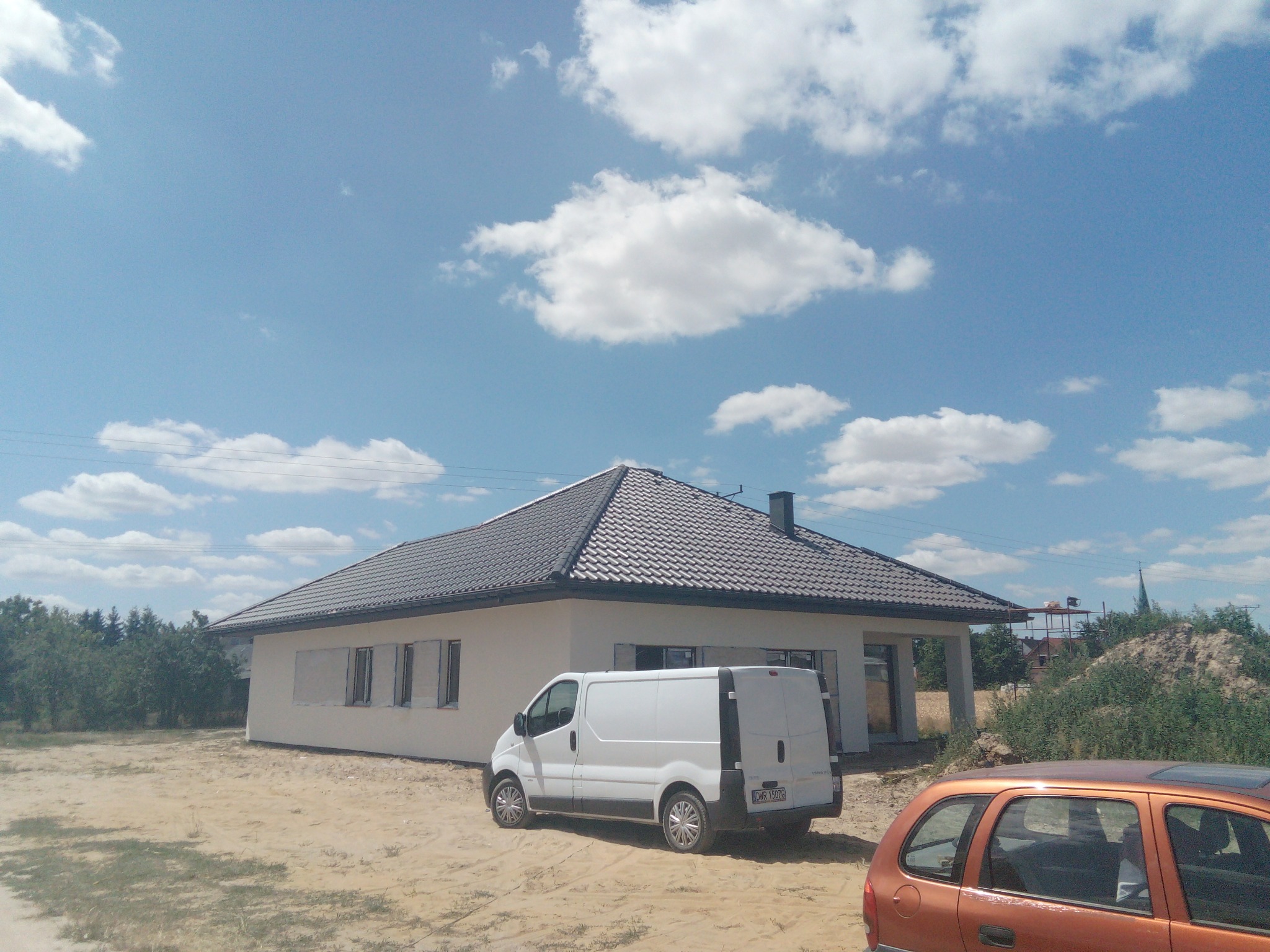 Nowy, ciemnoszary dach z dachówki ceramicznej na parterowym domu z białym tynkiem, zaparkowany biały van i pomarańczowe auto osobowe na piaszczystym terenie, błękitne niebo z chmurami w tle.