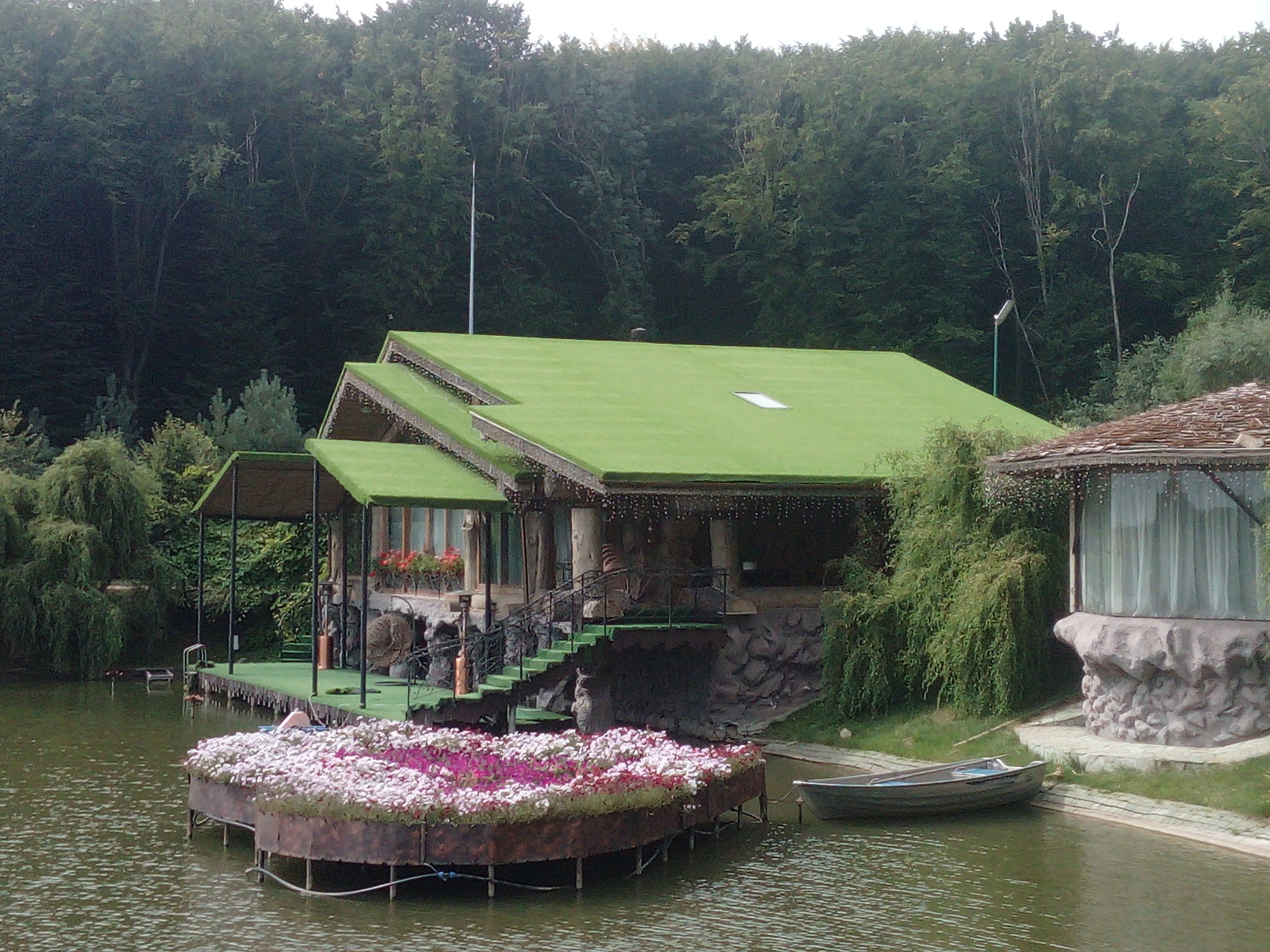 Dom z zielonym dachem nad jeziorem, z pływającą platformą z kwiatami, łódką i schodami.