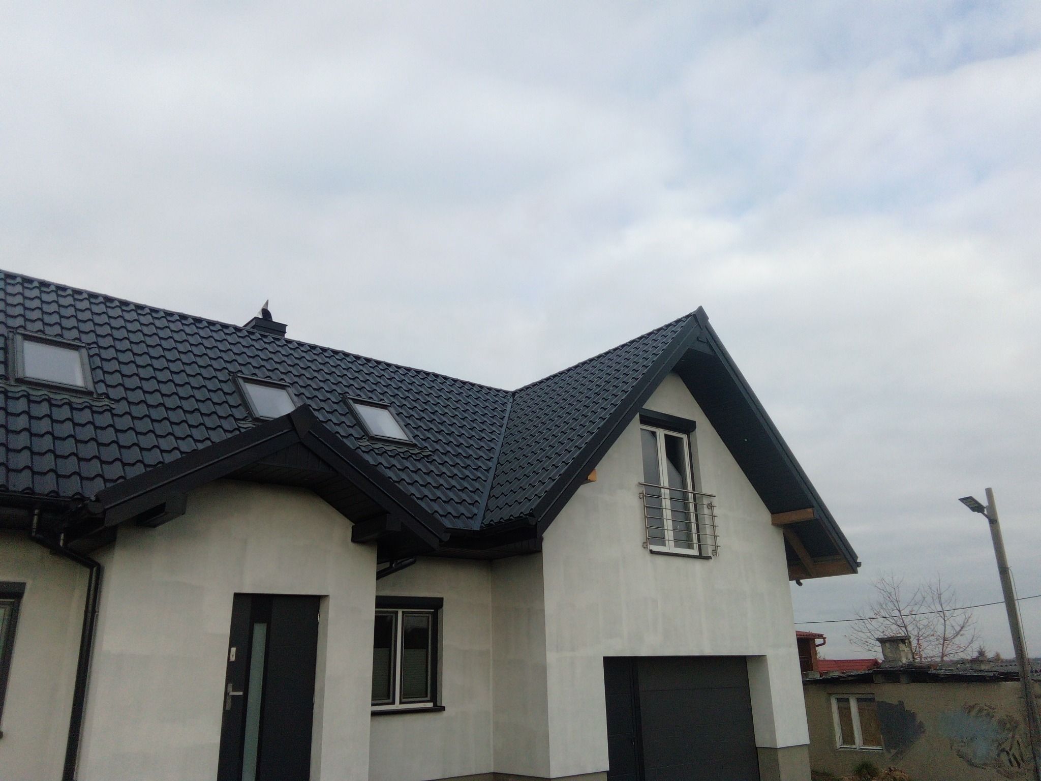 Nowoczesny dom jednorodzinny z ciemną dachówką ceramiczną, białymi ścianami i czarnymi detalami, widok z dołu na tle pochmurnego nieba.