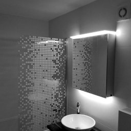 Remart - Czarno-białe zdjęcie wykończonej łazienki z kabiną prysznicową z wzorem pikseli, owalną umywalką na ciemnym blacie, lustrem z podświetleniem LED i wentylatorem w rogu.