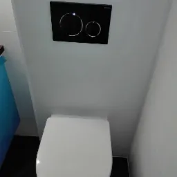 Nowoczesna toaleta podwieszana z czarnym panelem spłukującym Geberit na białych płytkach w wąskiej łazience.