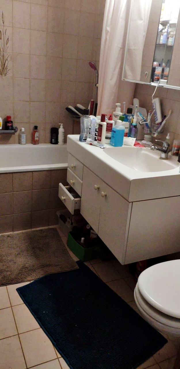 Umywalka z otwartą szufladą i licznymi kosmetykami w łazience z beżowymi płytkami i dywanikami.