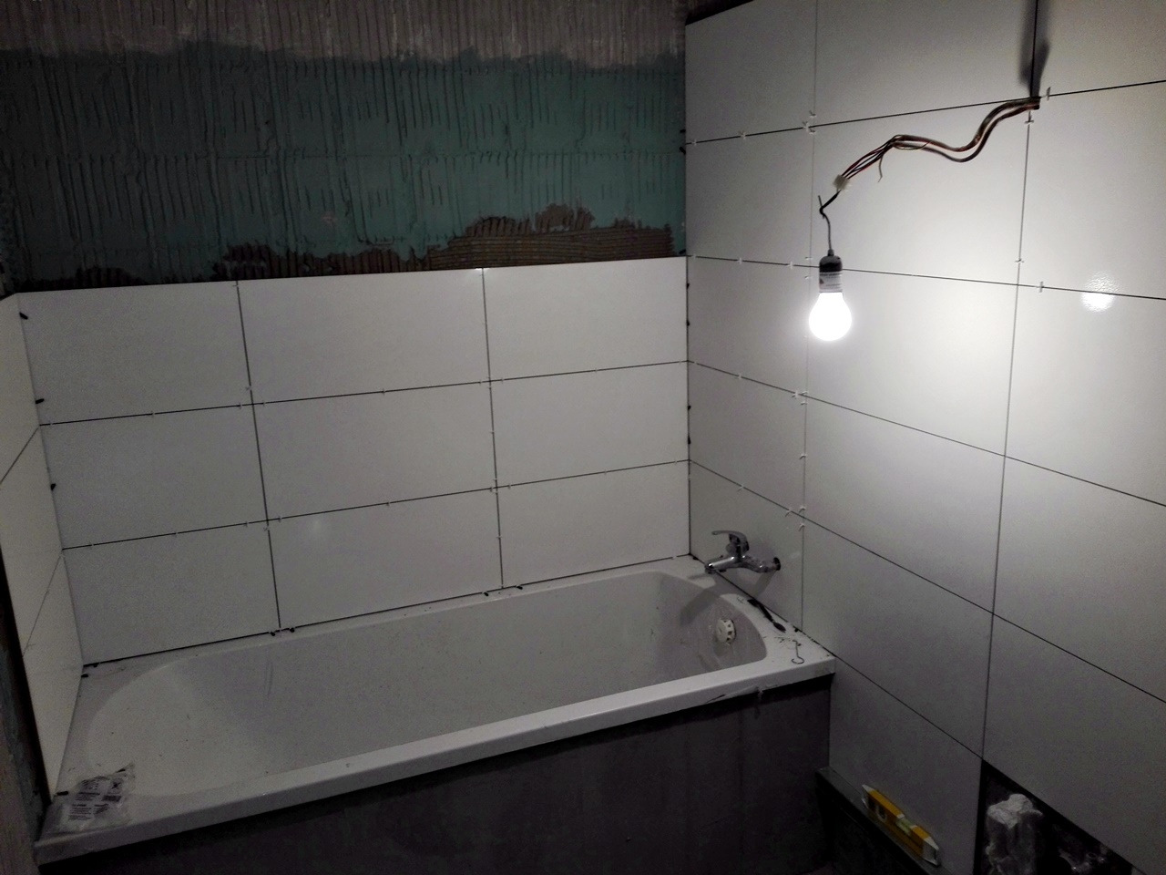 Łazienka w trakcie remontu: białe płytki ceramiczne układane na ścianach nad wanną, widoczne krzyżyki dystansowe, surowy fragment ściany z pozostałościami kleju i wisząca żarówka na kablu.