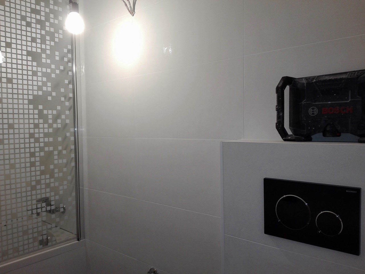 Nowoczesna łazienka z białymi płytkami, kabiną prysznicową z wzorem kwadratów, czarnym panelem sterującym spłuczką Geberit i radiem budowlanym Bosch na półce.