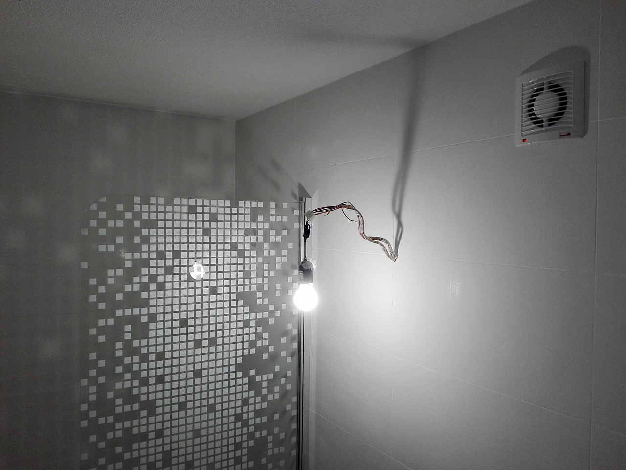Łazienka w trakcie wykończenia: widoczna kabina prysznicowa z mozaiką, wentylator na ścianie i tymczasowe oświetlenie z wiszącą żarówką oraz wystające przewody elektryczne.