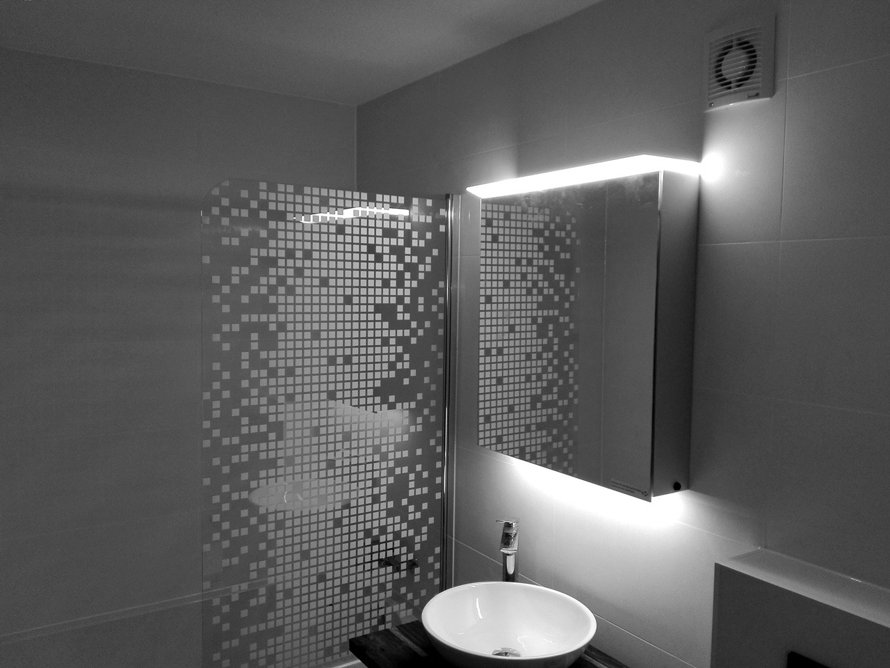 Czarno-białe zdjęcie wykończonej łazienki z kabiną prysznicową z wzorem pikseli, owalną umywalką na ciemnym blacie, lustrem z podświetleniem LED i wentylatorem w rogu.