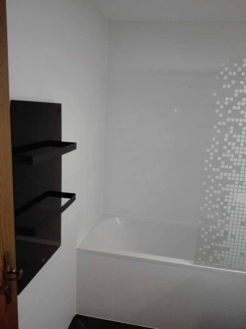 Minimalistyczna łazienka z białą wanną, czarną półką i mozaikową ścianą w odcieniach szarości.