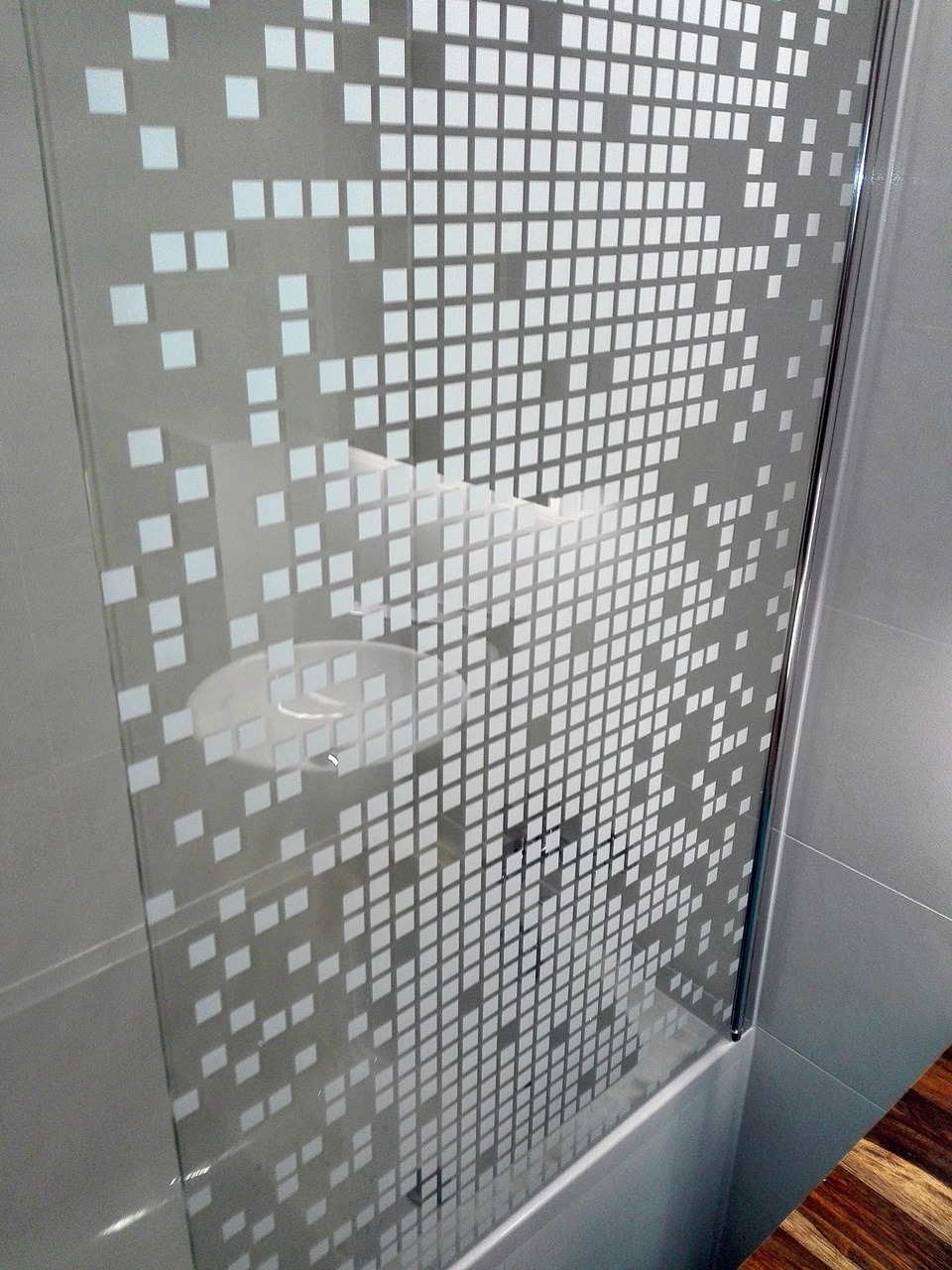 Szklana kabina prysznicowa z dekoracyjną folią w geometryczny wzór pikseli, widoczna umywalka w tle.