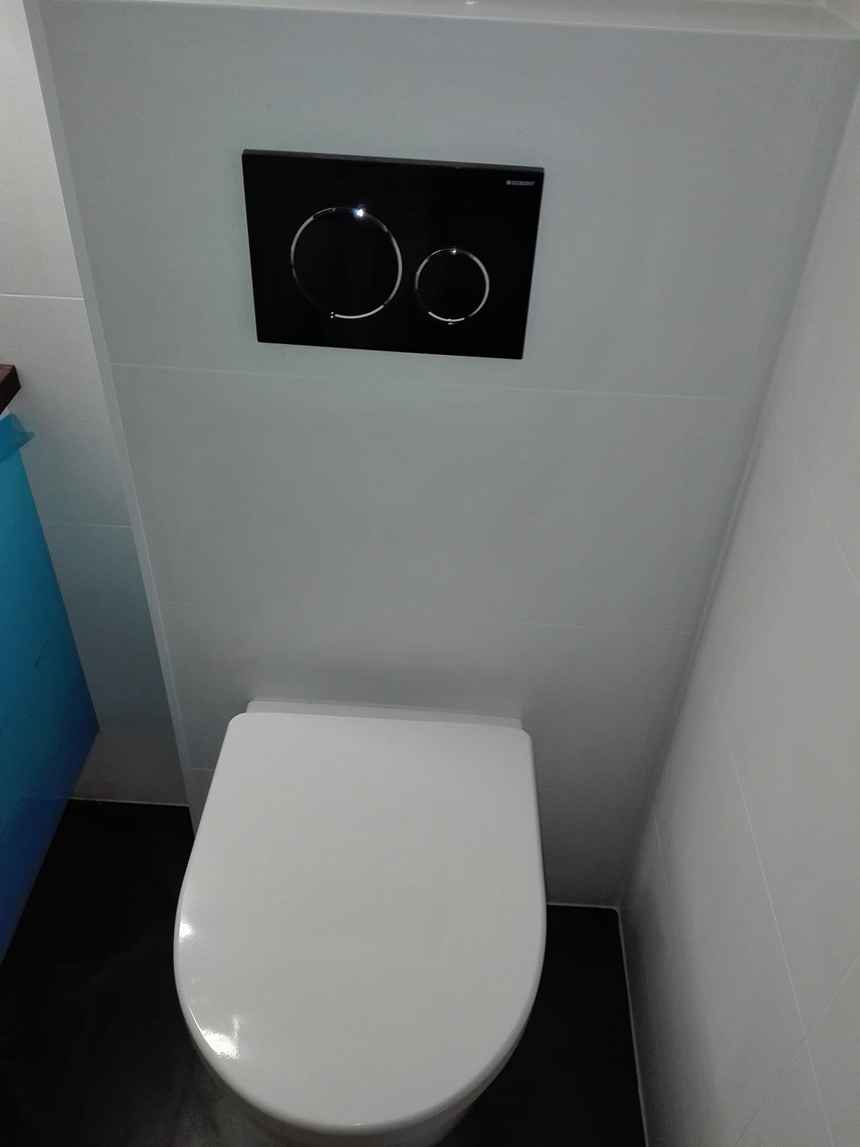 Nowoczesna toaleta podwieszana z czarnym panelem spłukującym Geberit na białych płytkach w wąskiej łazience.