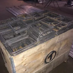 Kwadratowe elementy zbrojeniowe ułożone warstwami w drewnianej skrzyni z czarnym logo na boku, w tle widoczne długie pręty.