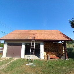 LUXPURE - Częściowo wyczyszczony, pomarańczowy dach z dachówki ceramicznej, drabina oparta o budynek gospodarczy z otwartym garażem i składowanym drewnem, widoczne ślady czyszczenia.